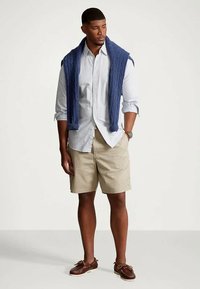 Άνδρας που φοράει ανοιχτόχρωμο καρό πουκάμισο με κουμπιά, μπεζ σορτς, ναυτικό πουλόβερ πλεξίματος ριπ draped στους ώμους και καφέ παπούτσια σκάφους, όρθιος.