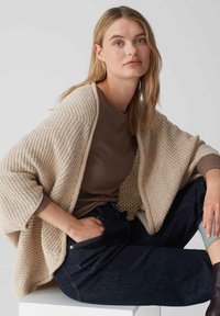 Gilet en tricot beige avec un motif texturé sur un haut à côtes marron, associé à un jean en denim foncé. Modèle assis dans une pose décontractée.