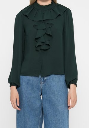 Blouse vert foncé à manches longues avec col volanté et détails sur le devant, portée avec un jean bleu large sur une personne.