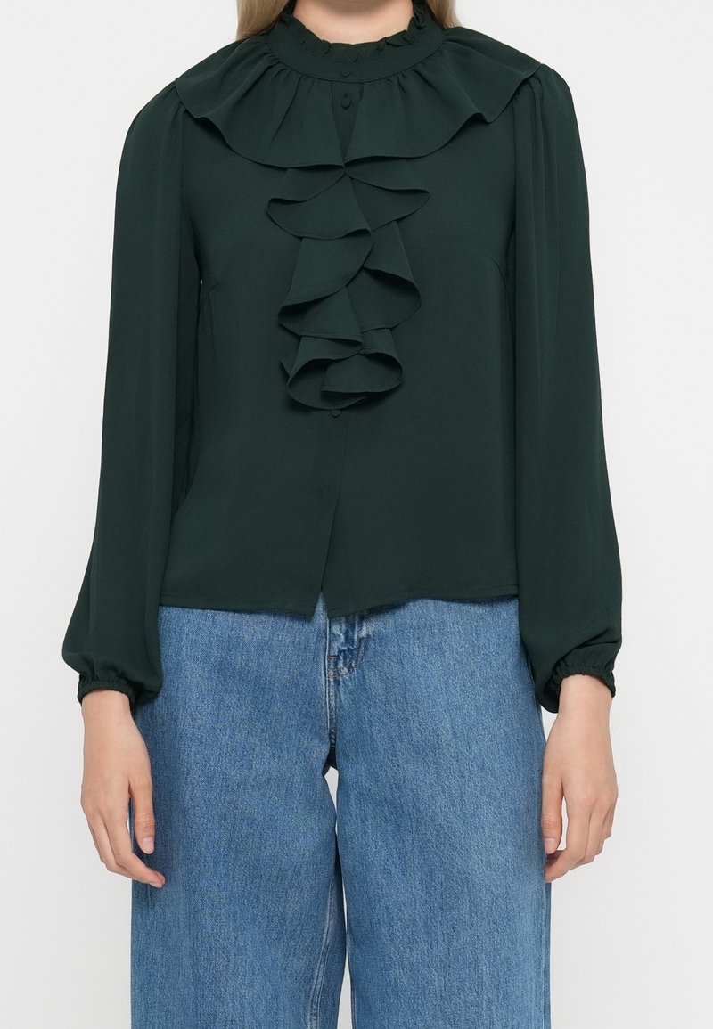 Blouse vert foncé à manches longues avec col volanté et détails sur le devant, portée avec un jean bleu large sur une personne.