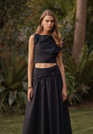 Fiatal nő hosszú hajjal, fekete ujjatlan crop topot és magas derekú fekete szoknyát visel, kertben állva.