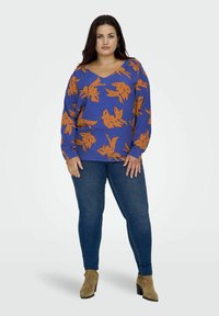 Blaues Langarmshirt mit orangefarbenem Blättermuster, V-Ausschnitt-Design, kombiniert mit dunklen Jeans und hellbraunen Stiefeletten. Glatte Textur, lockere Passform.