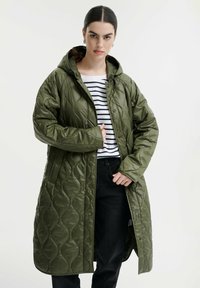 Cappotto lungo imbottito verde con cappucci, dotato di chiusure a scatto e una texture lucida, indossato sopra una maglietta a righe e pantaloni neri.