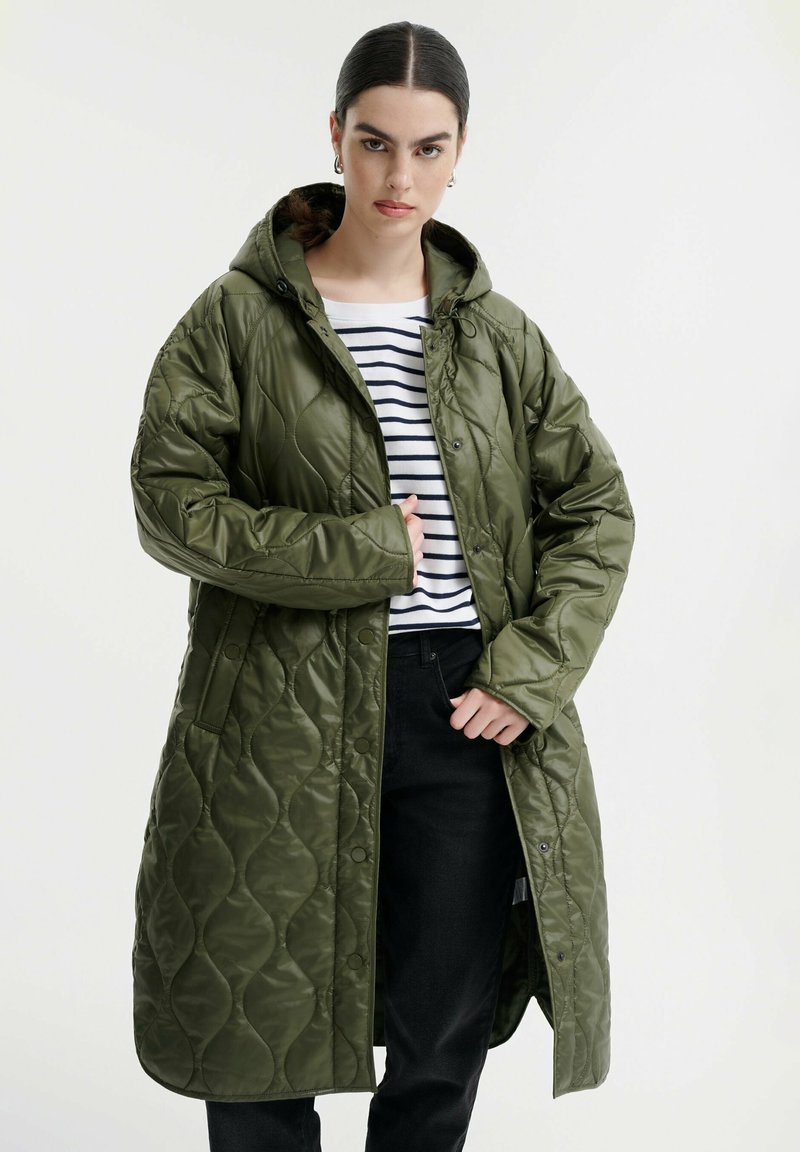 Cappotto lungo imbottito verde con cappucci, dotato di chiusure a scatto e una texture lucida, indossato sopra una maglietta a righe e pantaloni neri.