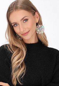 Runde, farbenfrohe Ohrringe mit floralen Mustern in pastellfarbenen und metallischen Akzenten, getragen von einer Person in einem schwarzen Pullover.