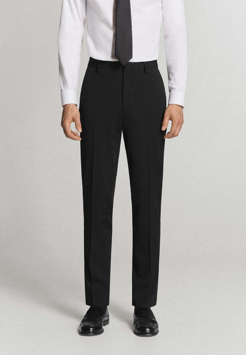 Homme portant un pantalon de costume noir, des chaussures en cuir noires, une chemise blanche et une cravate noire, debout devant un fond clair.