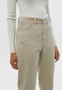 Beige corduroy broek met verticale ribbels, hoge taille, een enkele voorkant- en achterkantzak en metalen knoopaccenten. Gekleed met een aansluitend wit shirt.