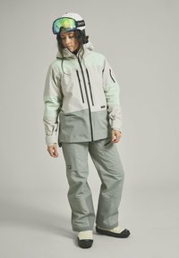 Snow jacket in light gray and mint green, with multiple zip pockets and adjustable cuffs. Paired with matching gray snow pants and black boots.