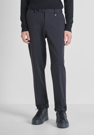 Hombre vestido con pantalones negros a medida y zapatillas negras con cordones de pie contra un fondo gris claro.