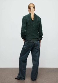 Suéter verde de punto con patrón de trenzas, combinado con jeans de mezclilla azul sueltos. Vista trasera; presenta una abertura de pierna ancha y dos bolsillos traseros.
