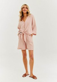 Svetloružová ľanová košeľa zviazaná v páse, doplnená o zodpovedajúce šortky. Oblečenie má voľný strih a textúrovaný povrch, doplnené hnedými sandálmi.
