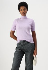 Lillā īsām piedurknēm turtlenecks džemperis no mīksta auduma, apvienots ar augsto jostu tumši pelēkiem džinsiem un turot nelielu balto somiņu.