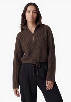 Femme portant un pull en maille côtelée marron avec une fermeture éclair quart et un ourlet à cordon, associé à un pantalon noir ample, debout devant un fond blanc.