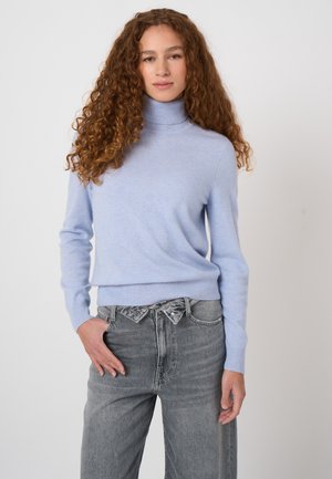Vrouw met lang krullend haar die een lichtblauwe coltrui en hooggetailleerde grijze spijkerbroek draagt, staand tegen een effen achtergrond.