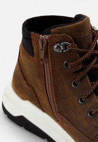 Stivale alto in suede marrone con accenti neri, dotato di zip laterale, lacci marroni e suola in gomma testurizzata per una migliore aderenza.
