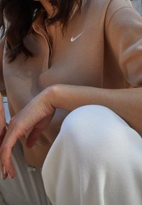 Žena v béžovém dlouhém rukávu Nike s bílým logem, podpírající ruku na pokrčeném koleni v bílých kalhotách, s viditelnými vlnitými tmavými vlasy.