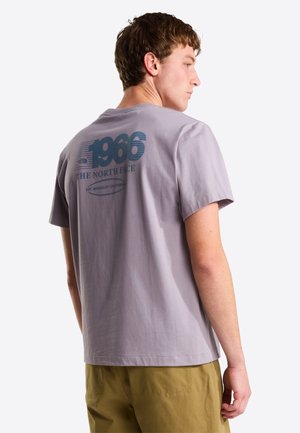 Jeune homme de profil portant un t-shirt lavande avec le texte "1966 The North Face" et un pantalon kaki, sur un fond blanc uni.