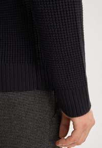 Zbliżenie na dłoń spoczywającą obok mankietu czarnego, teksturowanego swetra z dzianiny oraz szarych, w kratę spodni.