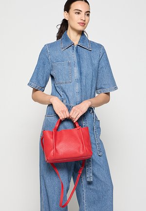 Woman wearing a short-sleeve denim jumpsuit holding a red leather handbag with dual handles and a shoulder strap.
