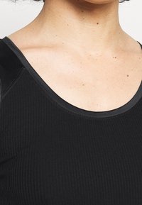 Schwarzes, geripptes Tanktop mit rundem Ausschnitt und glattem, kontrastierenden schwarzen Saum entlang der Kanten. Leicht taillierte Passform.