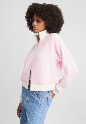 Veste zippée rose clair avec texture côtelée, rayures blanches et col. Présente des poches latérales et est assortie à un jean rayé bleu.