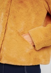 Handen delvis i fickan på en senapsgul fuskpälsjacka som bärs över mörkblå byxor med vit sömnad.