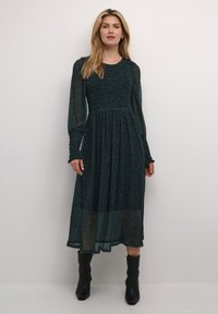 Pitkä, tumma teal-mekko, jossa on rypistetty yläosa, läpikuultavat pitkät hihat ja ylivoimaisesti laskeutuva hame. Yhdistettynä mustiin nilkkasaappaisiin. Rakenteellinen materiaali, jossa on hillitty kuvio.