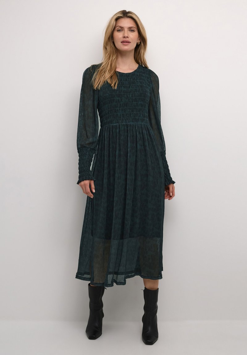 Pitkä, tumma teal-mekko, jossa on rypistetty yläosa, läpikuultavat pitkät hihat ja ylivoimaisesti laskeutuva hame. Yhdistettynä mustiin nilkkasaappaisiin. Rakenteellinen materiaali, jossa on hillitty kuvio.