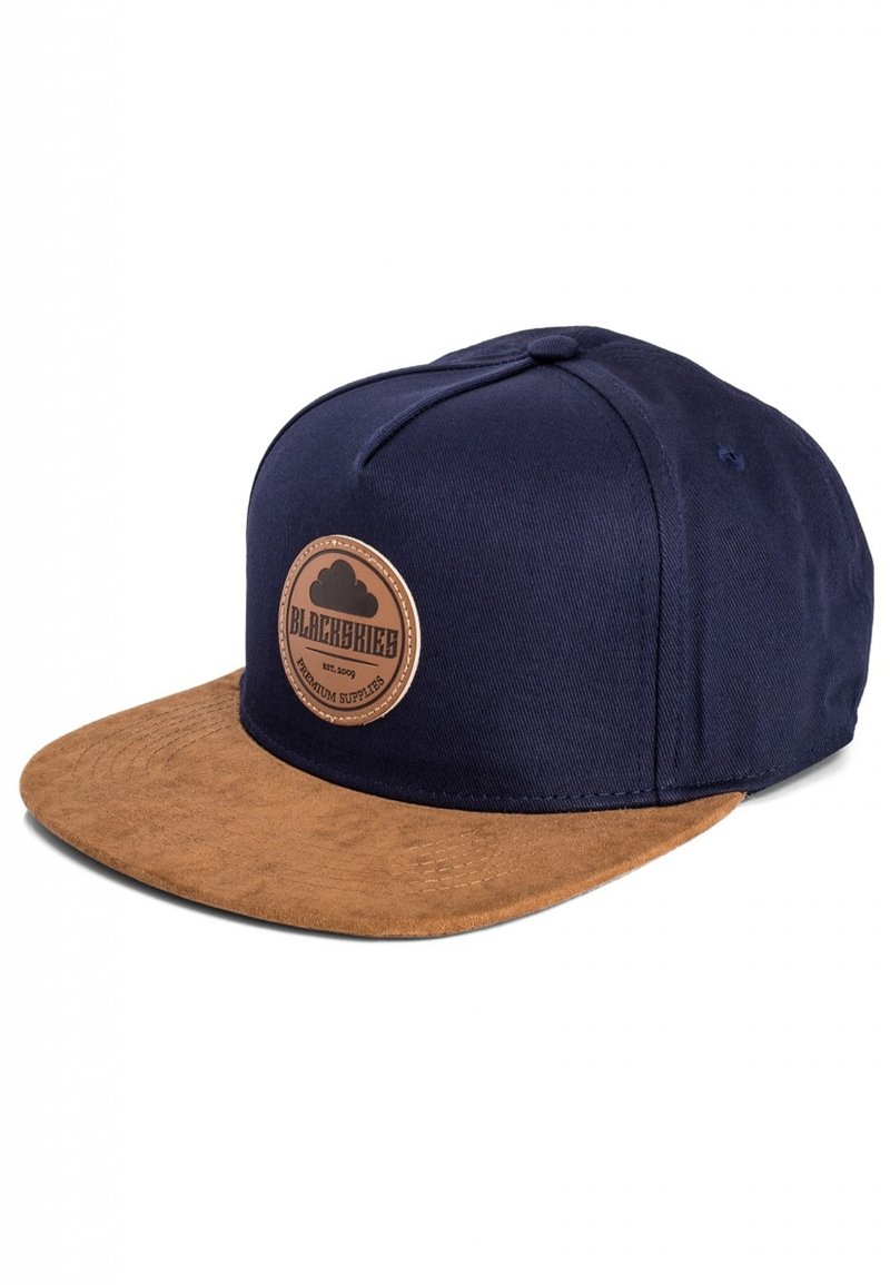 Navy blue cap with a brown suede brim, featuring a circular leather patch with text. Six-panel design and button top detail.