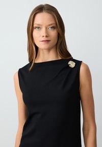 Top senza maniche nero realizzato in tessuto liscio, con scollatura rotonda e un fermaglio metallico testurizzato sulla spalla sinistra.