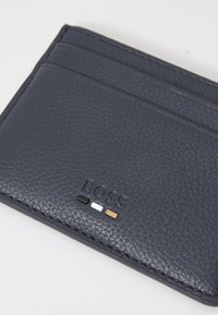 Tamsiai mėlynas odinis kortelių laikiklis su tekstūruota paviršiumi, turintis kelis kortelių lizdus ir įspaustą "BOSS" logotipą su juodais, baltais ir rudais akcentais.