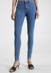 Jeans ajustados de mezclilla azul de talle alto con una textura suave; cuentan con trabillas para el cinturón, cremallera frontal y dos bolsillos traseros. Combinados con mule negros de tacón.