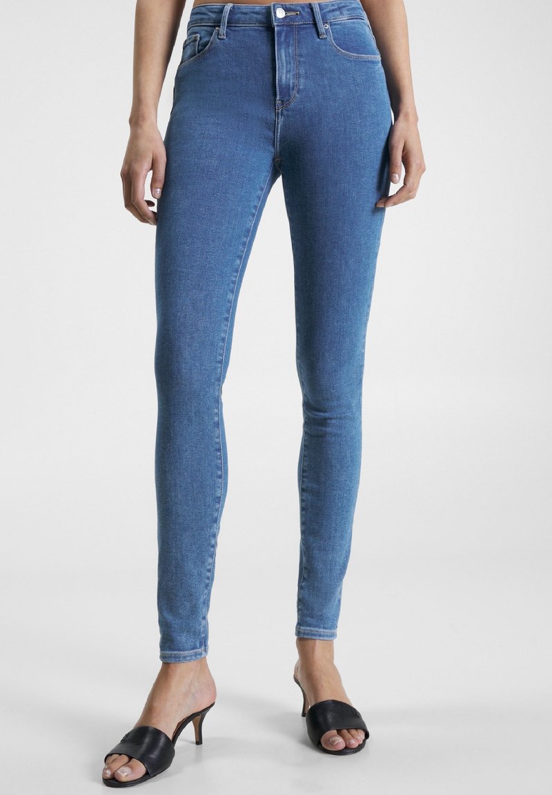 Jeans ajustados de mezclilla azul de talle alto con una textura suave; cuentan con trabillas para el cinturón, cremallera frontal y dos bolsillos traseros. Combinados con mule negros de tacón.