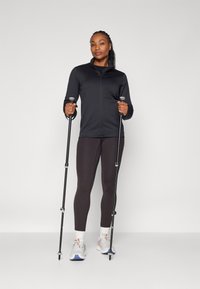 Zwarte zip-up jas met hoge kraag, zwarte leggings en wandelstokken. Grijze sneakers met oranje accenten. Gladde textuur overal.