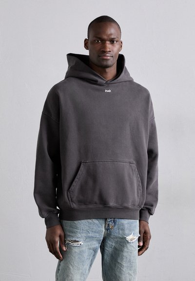 Grijze katoenen hoodie met een voorzak en geribbelde manchetten. Heeft een logo op de borst. Draagt over versleten lichtblauwe jeans.