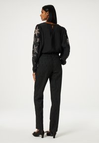 Zwarte blouse met geborduurde sterdetails op de mouwen, gecombineerd met zwarte broek met streepjes en zwarte hoge hakken. Sleutelgat aan de achterkant.