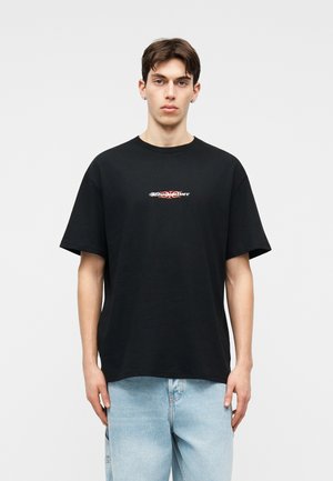 Mladi moški, oblečen v črno preveliko majico z majhnim belim in rdečim napisom na prsih ter svetlo modrimi kavbojkami, stoji ob preprostem ozadju.