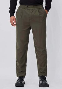Pantalon vert olive avec plis, une taille �élastique confortable, deux poches avant et une coupe fuselée, associé à des chaussures noires à lacets.