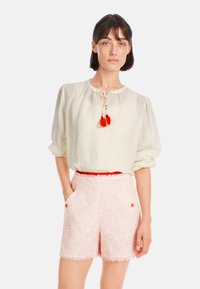 Helles gelb, locker sitzende Bluse mit gerafftem Ausschnitt und roten Quasten; kombiniert mit rosa strukturierten Shorts mit fransigen Kanten und Taschen.