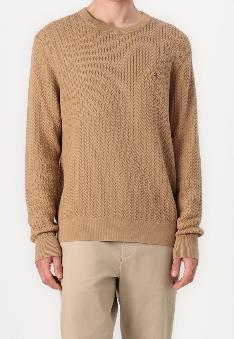 Pull beige avec un motif chevrons texturé, col rond et poignets côtelés. Petit logo sur la poitrine, porté avec un pantalon clair.