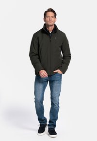 Donkergroene jas met een hoge kraag, ritssluiting op de borst en zijzakken. Gecombineerd met blauwe jeans en zwarte schoenen. Eenvoudig, functioneel ontwerp.