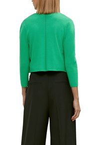 Mujer vestida con un suéter cortado de color verde brillante y mangas largas, y pantalones anchos negros, vista desde atrás.