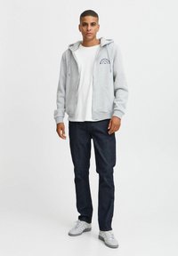 Lys grå hettegenser med glidelås og frontlomme, med et marineblått grafisk design på brystet. Kombinert med mørk denim jeans og grå joggesko.