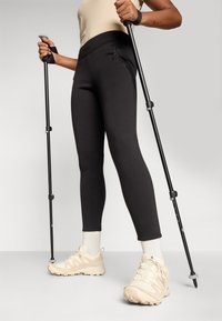Svarta träningsleggings med hög midja, i kombination med beige vandringsskor. Användaren håller två vandringsstavar, vilket visar på funktionalitet och grepp.
