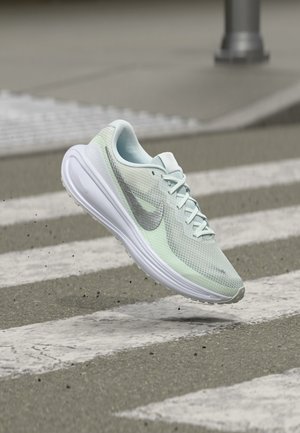 Chaussure de course Nike vert clair et blanc en l'air au-dessus d'un passage piéton texturé sur l'asphalte.