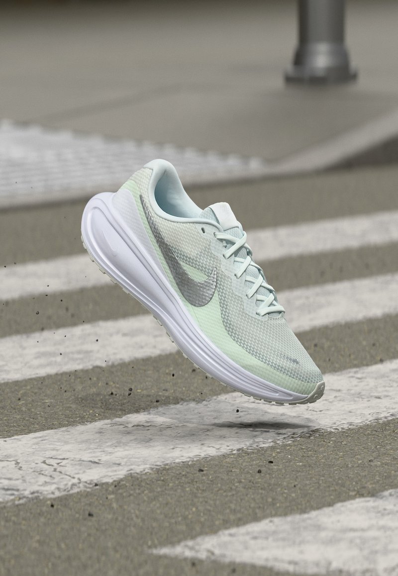 Ανοιχτό πράσινο και λευκό παπούτσι Nike για τρέξιμο στον αέρα πάνω από μια υφασμένη διάβαση πεζών σε άσφαλτο.