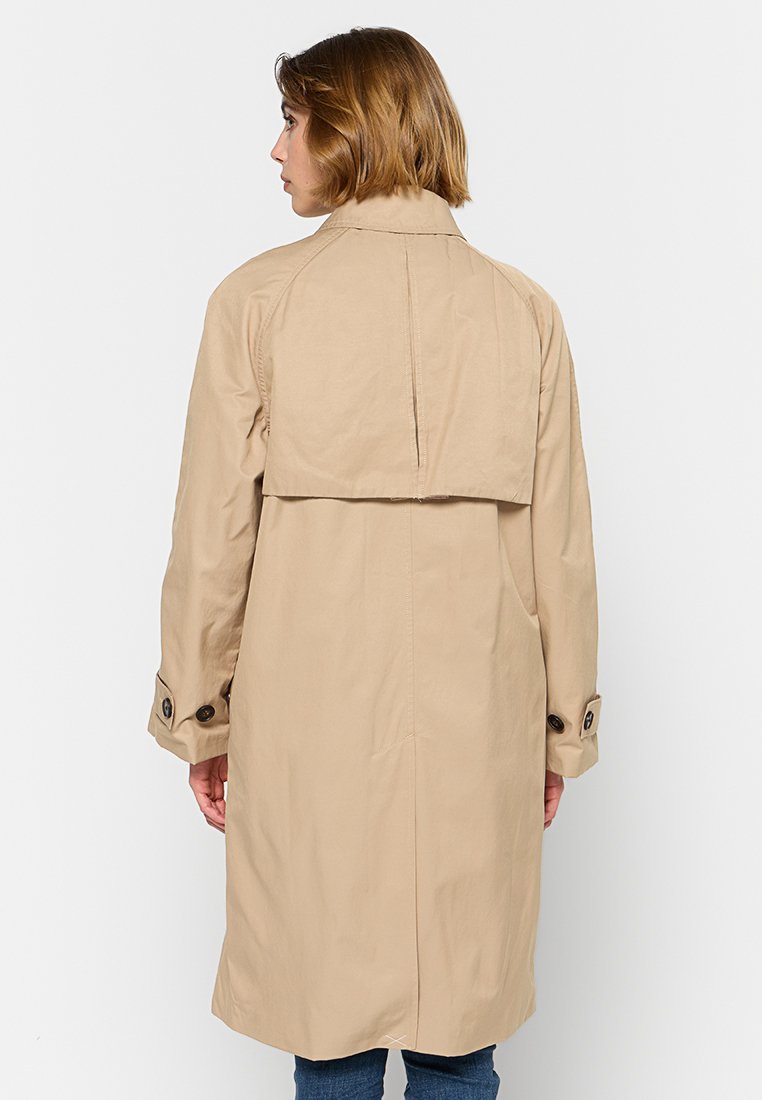 Cappotto trench beige, di lunghezza media, realizzato in tessuto liscio. Presenta una pattina sul retro, polsini con bottoni e due tasche laterali.