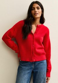 Rode cardigan van zacht materiaal, met een diepe V-hals en knoopsluiting. Lange mouwen en een aansluitende zoom, gecombineerd met blauwe jeans.