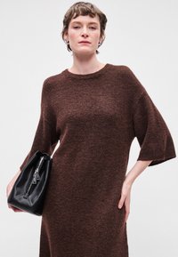 Brūna adīta kleita ar apaļu kakla izgriezumu un platu 3/4 piedurkni. Modelis tur nelielu melnu ādas somu ar sudraba rāvējslēdzēja detaļu.