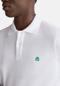 Camiseta polo blanca hecha de tela texturizada. Presenta un cuello abotonado y un pequeño logo de pulpo bordado en verde en el lado izquierdo del pecho.
